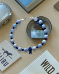 A beaded necklace with light blue and white marbled beads, deep blue beads, and a small blue fish charm, arranged on a beige tabletop with books and small decorative objects.