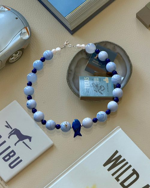 A beaded necklace with light blue and white marbled beads, deep blue beads, and a small blue fish charm, arranged on a beige tabletop with books and small decorative objects.